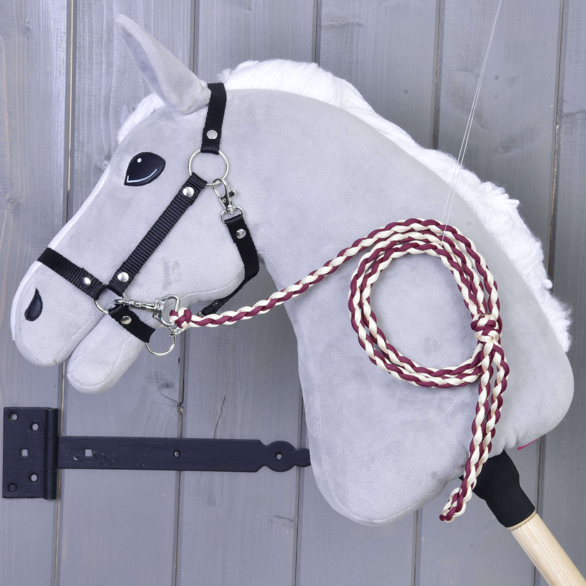 Bordó-krémszínű fonott vezetőszár egy szürke Hobby Horse ló fekete kötőfékére csatolva, szürke fafal mellett