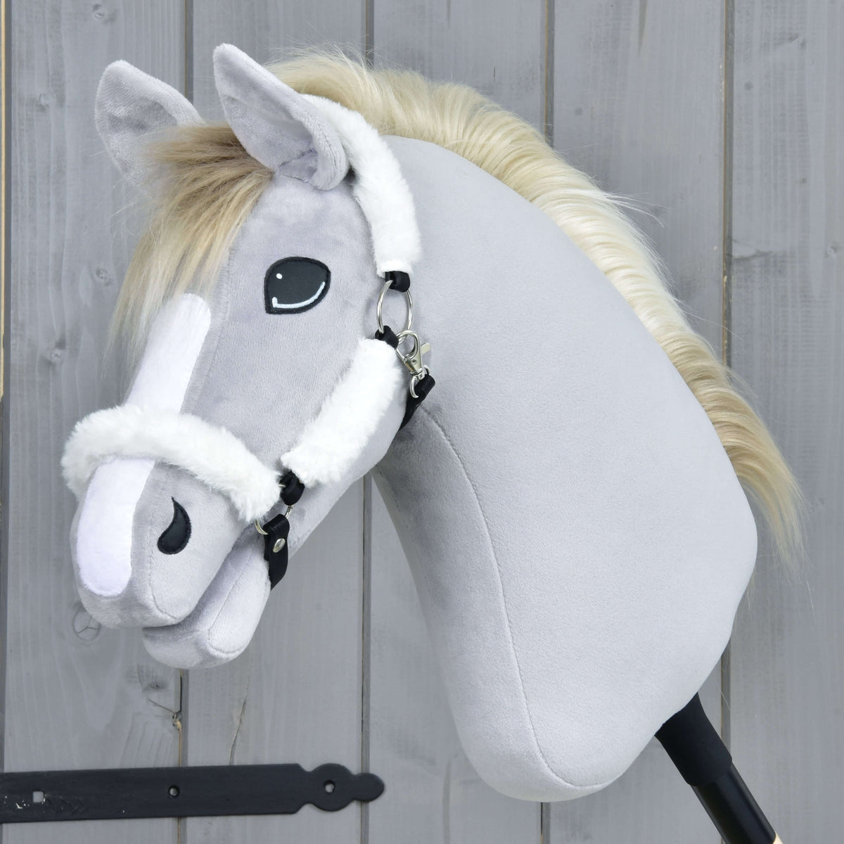 Szürke plüss Hobby Horse ló fehér hókával és krémszínű sörénnyel a fehér szőrmés kötőfékben, oldalnézetből, szürke fafal mellett