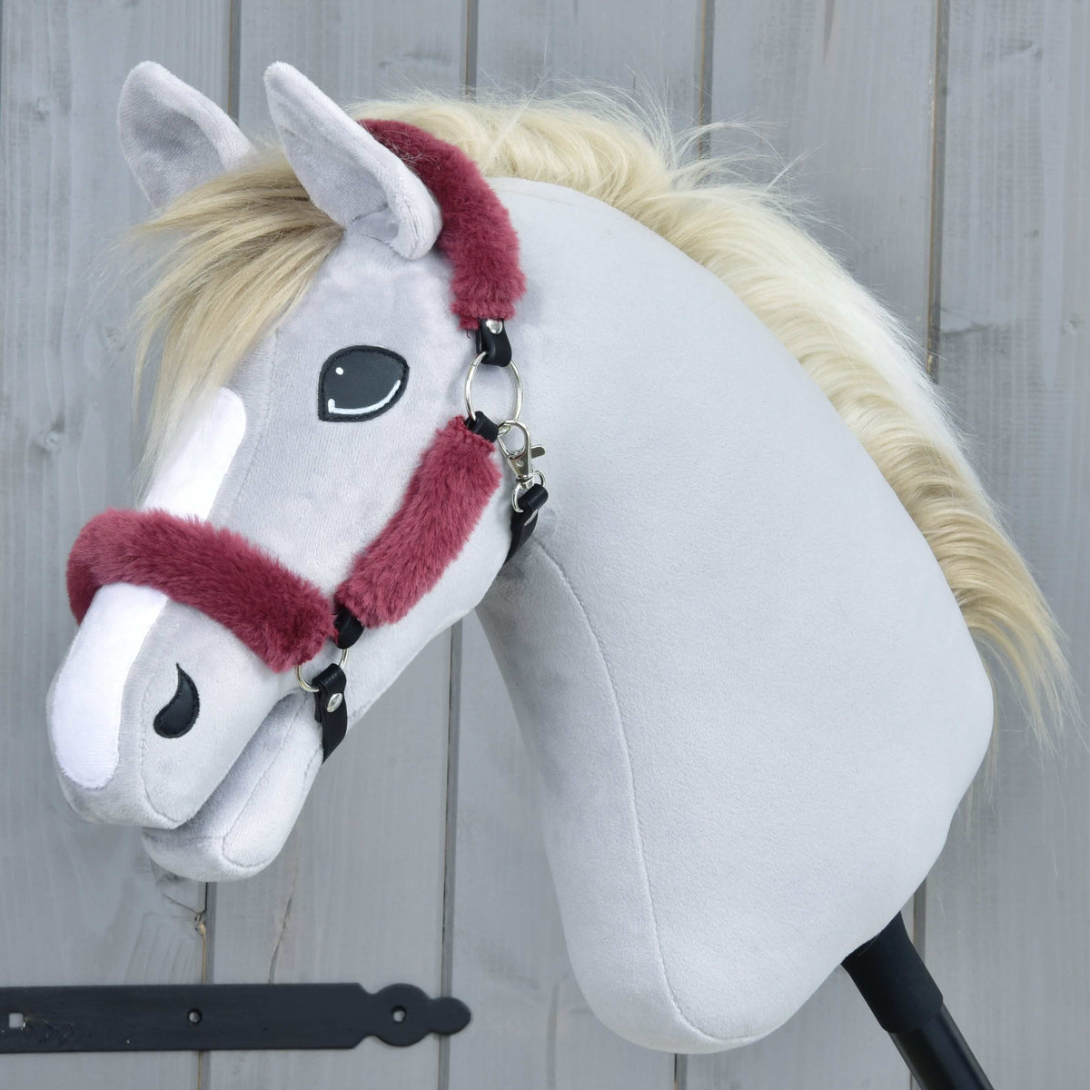 Szürke plüss Hobby Horse ló krémszínű sörénnyel a bordó szőrmés kötőfékben, oldalnézetből, szürke fafal előtt