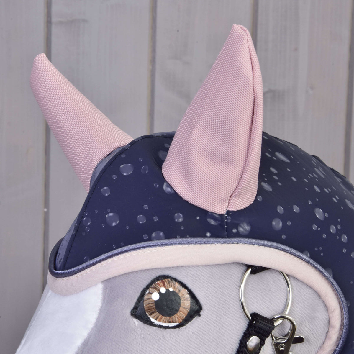 Close-up of the pale pink woven ears and blush trim of the Raincoat Pinkie Blue above the amber eye of a grey Hobby Horse