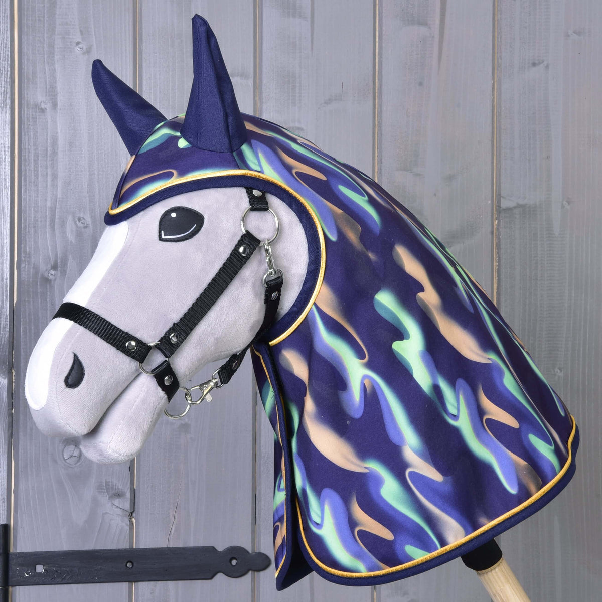Szürke Hobby Horse ló fekete kötőfékben, szivárványos esőtakaróban, amely a fejét és a nyakát is befedi, sötétkék fülekkel