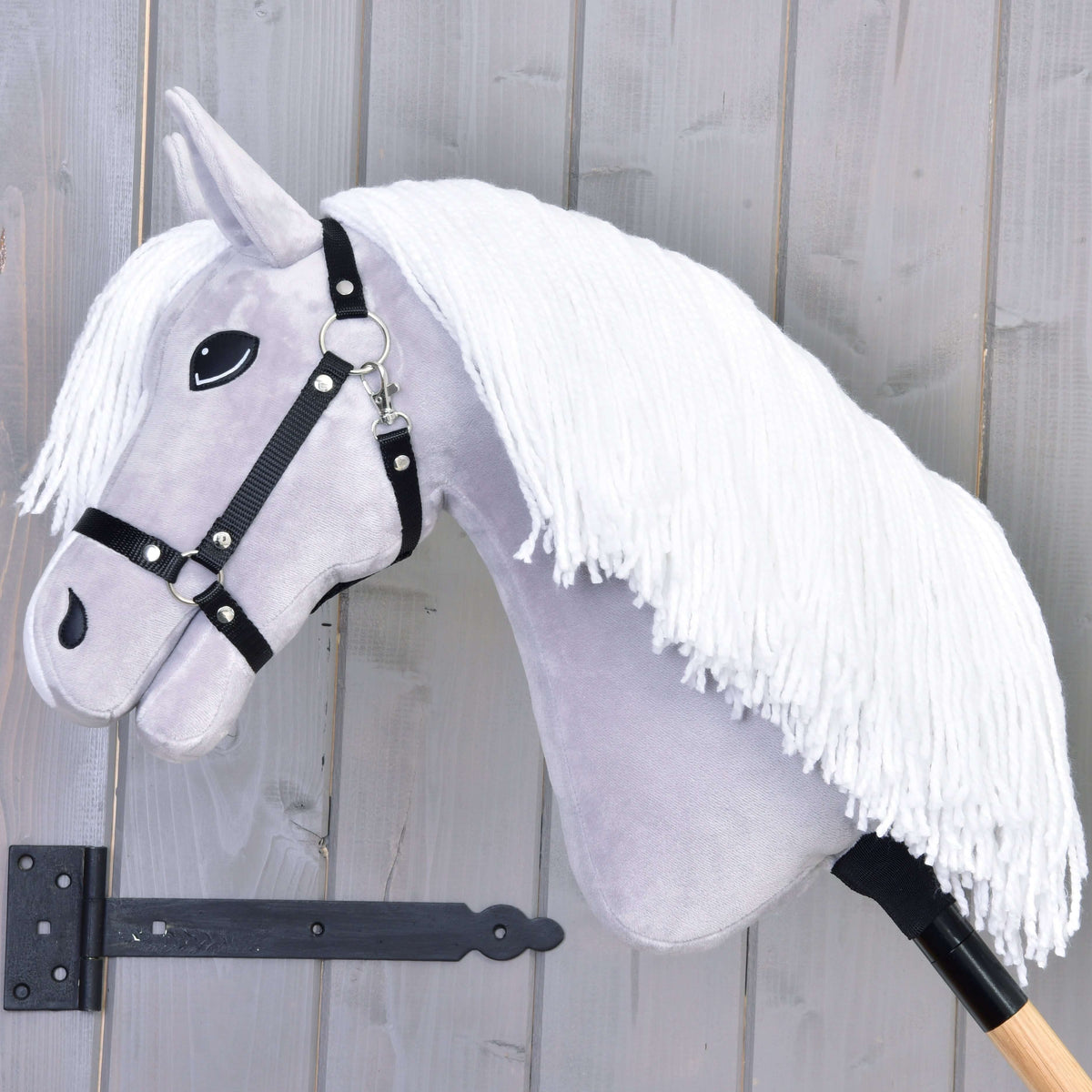 A világosszürke Hobby Horse ló oldalnézetből, hosszú fehér fonalsörénnyel és fekete kötőfékkel, faboton
