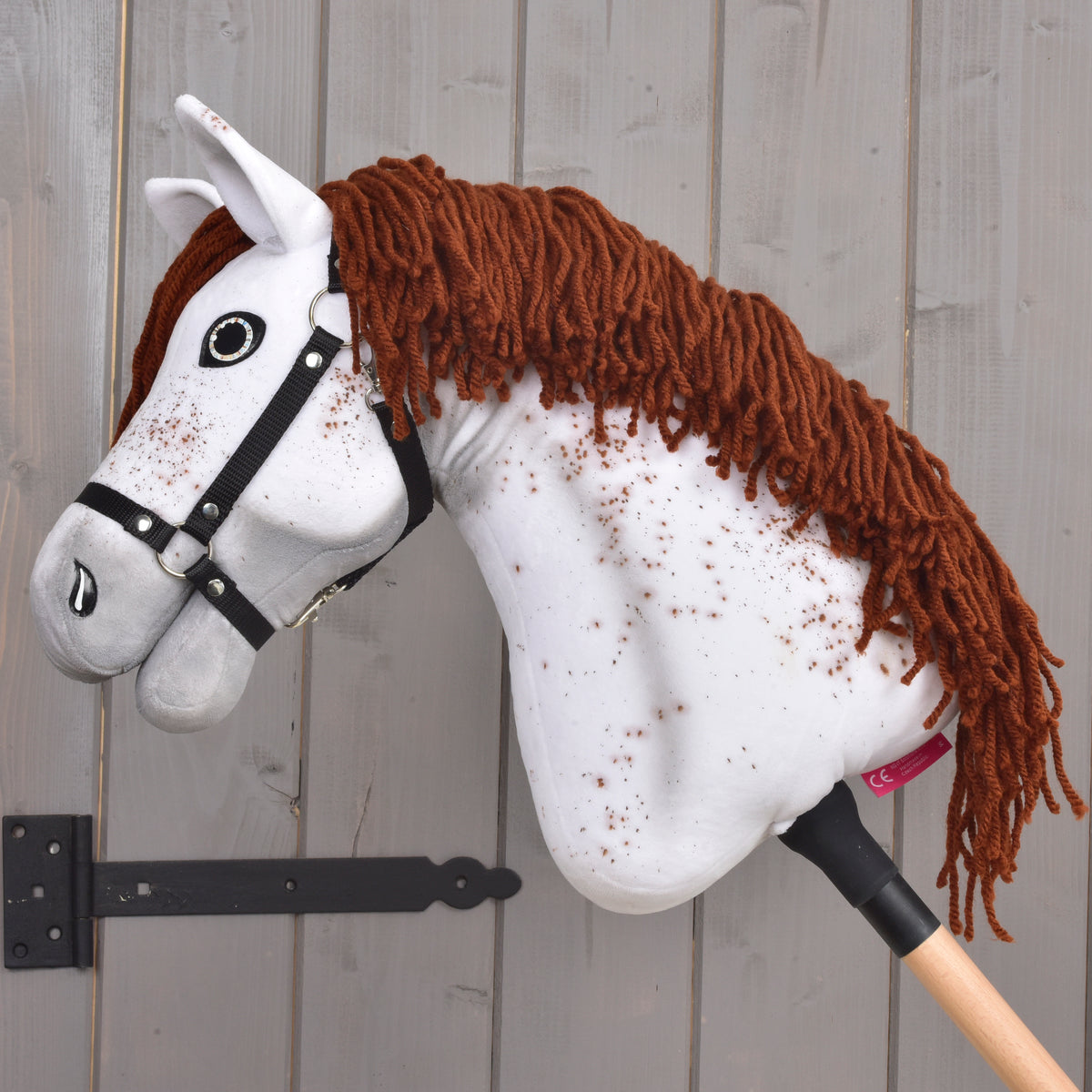Side view of the speckled Appaloosa Hobby Horse in a black halter with its long red-brown yarn mane against grey boards