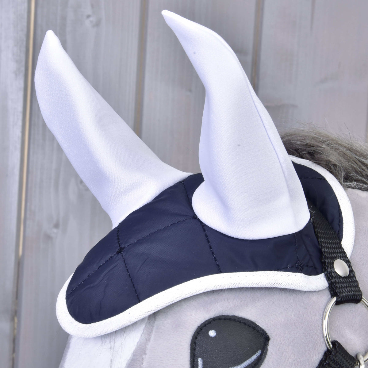 Közeli kép: a Monte szett fülvédője magas fehér fülekkel, sötétkék steppelt fejrésszel és fehér szegéssel egy szürke Hobby Horse lovon