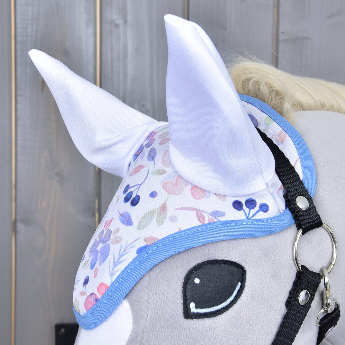 Közeli kép a virágmintás fülvédőről fényes fehér fülrészekkel és kék szegéllyel egy szürke Hobby Horse lovon, a fekete kötőfék mellett
