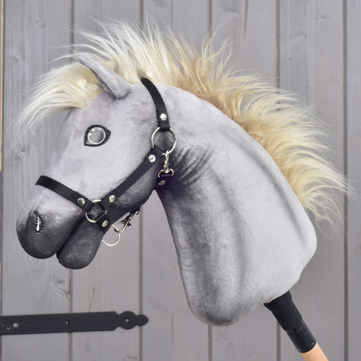 LarDen Hobby Horse Silver Dream szürke plüssfejjel, krémszínű sörénnyel és fekete kötőfékkel, oldalról, egy fa istállóajtó mellett