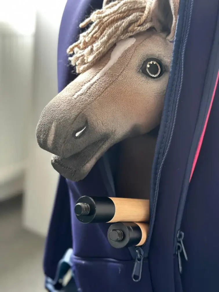 Bézs fonalsörényű, világosbarna Hobby Horse ló kukucskál ki egy sötétkék hátizsákból egy kétrészes fabot mellett