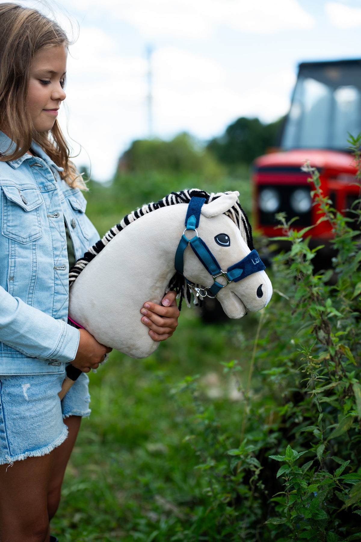 Farmerdzsekis lány kék kötőfékes Hobby Horse Aurora lovat tart a szabadban zöld növények mellett, mögötte piros traktor