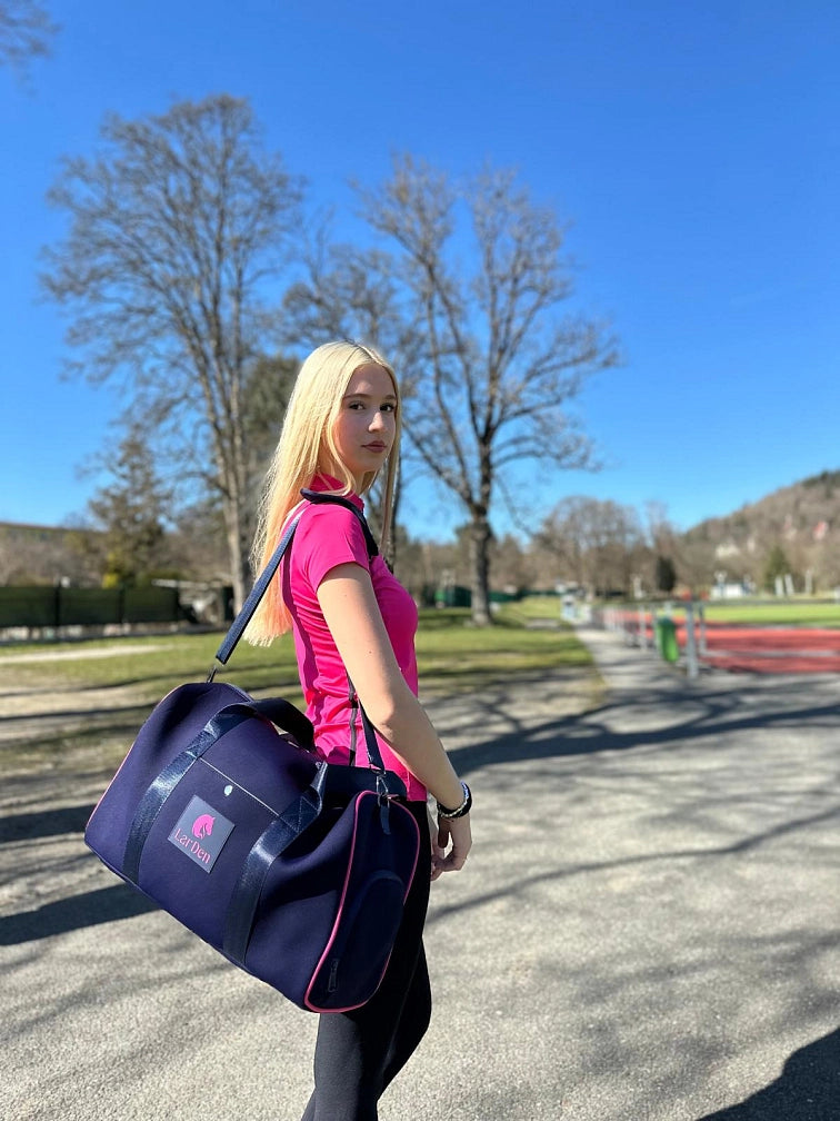 Hosszú szőke hajú fiatal nő rózsaszín sportpólóban, sötétkék sporttáskával áll a szabadban egy napsütéses napon a Hobby Horse lovával, készen egy vidám Hobby Horsing napra a tiszta kék ég és a lombos fák alatt.