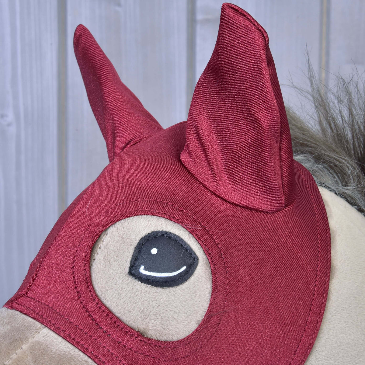 Részlet: a Barn Red maszk fényes sötétpiros fülrészei, alattuk tűzött szemnyílás, egy bézs Hobby Horse lovon