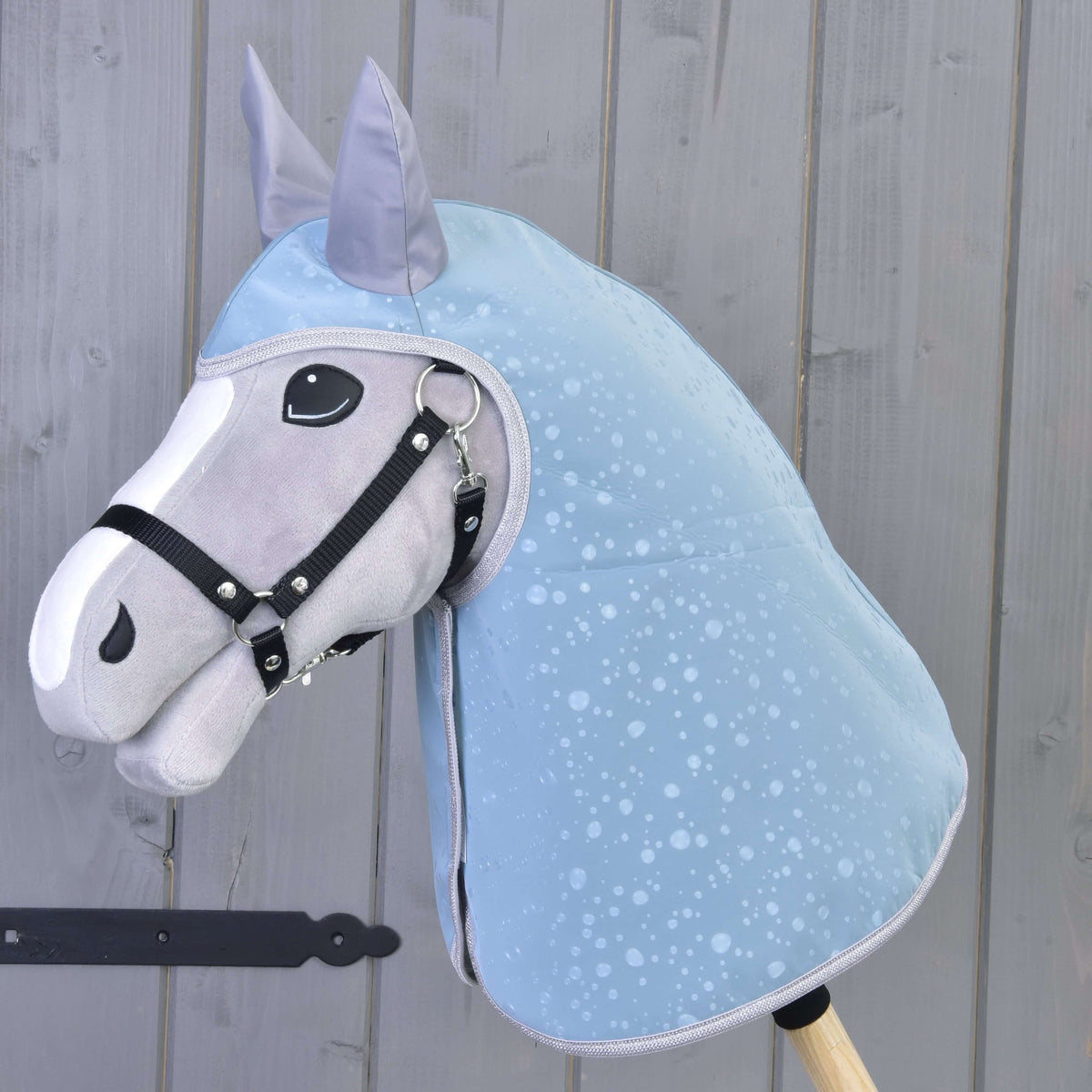 Esőtakaró Blue Drops egy fehér hókás, fekete kötőfékes szürke Hobby Horse lovon: a fejet és a nyakat is takarja, szürke deszkafal előtt
