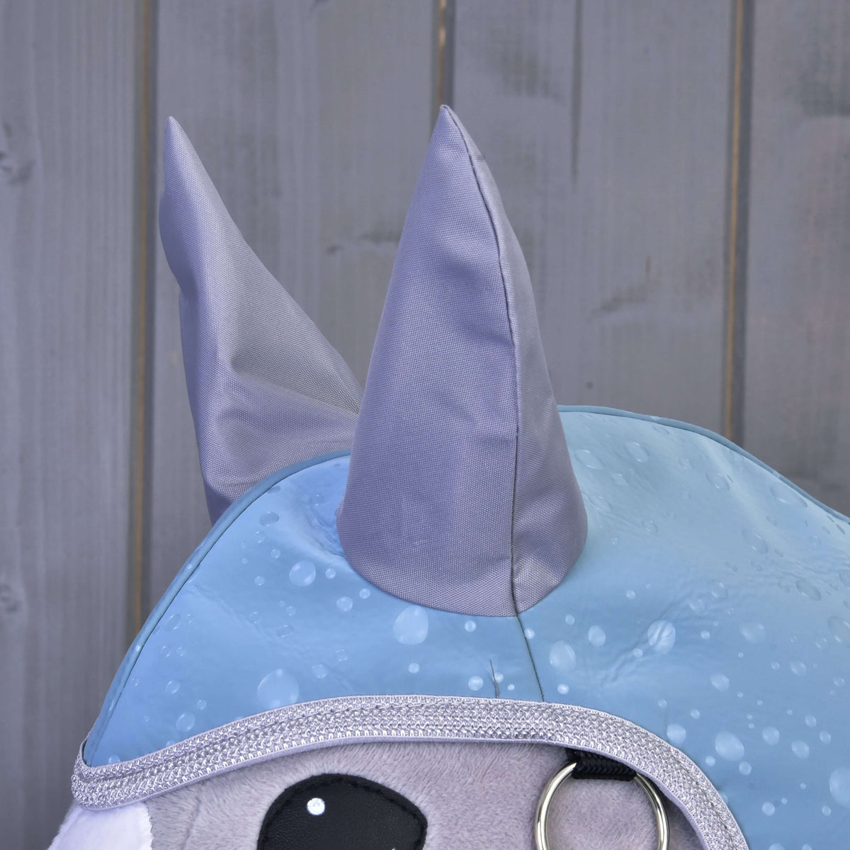 Közeli kép: a Blue Drops esőtakaró szürke szaténfülei és ezüstszínű csillogó szegélye egy szürke Hobby Horse lovon