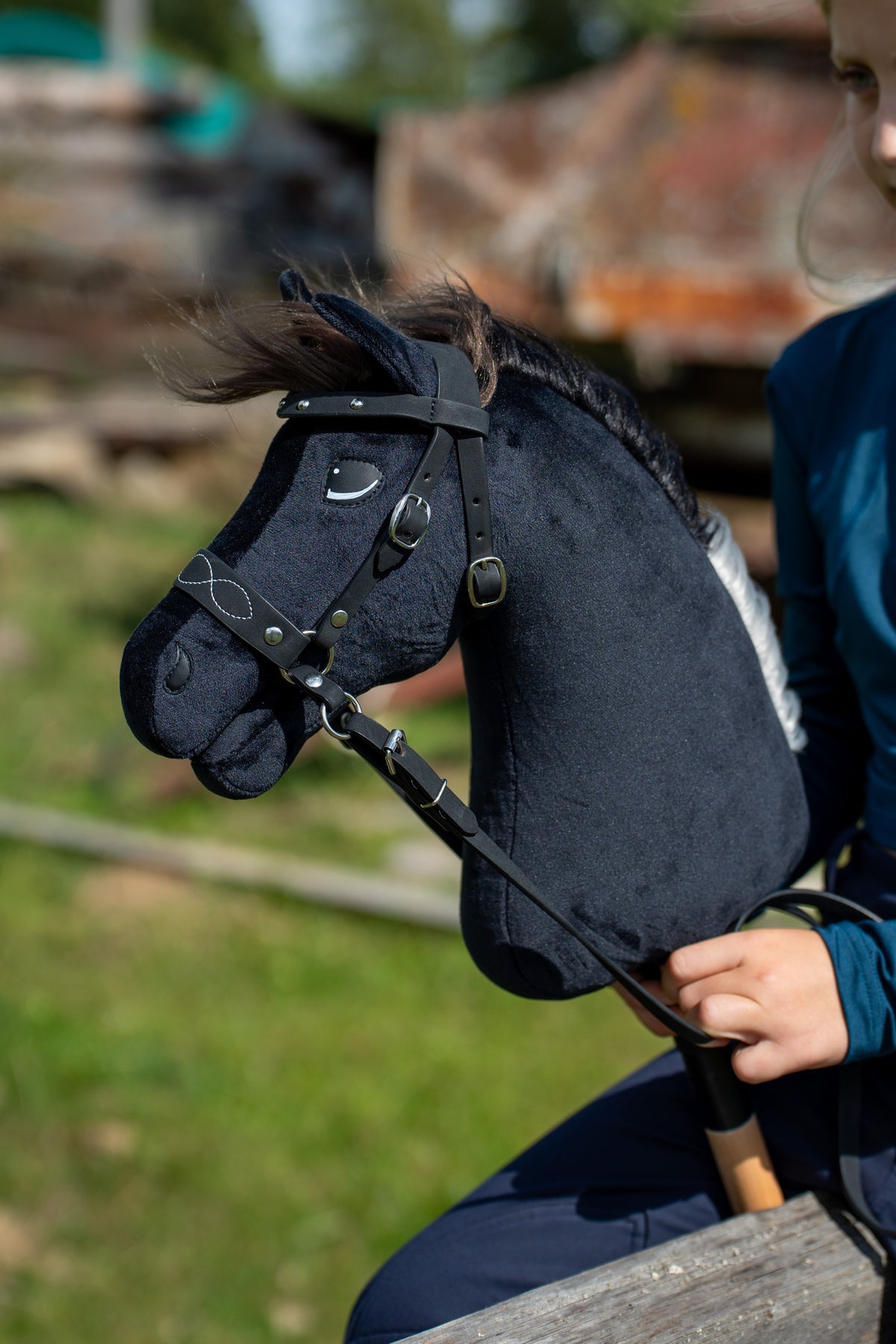 A fekete kötőfékkel kapható Hobby Horse Bond közelről, itt fekete kantárral, fehér tűzésű orrszíjjal és szárral, a háttérben elmosódott tanyaudvar