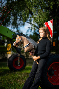 Fekete garbós lány zöld mezőgazdasági gépek mellett tartja a fekete kötőfékkel kapható Hobby Horse Bren lovat, itt fekete kantárral