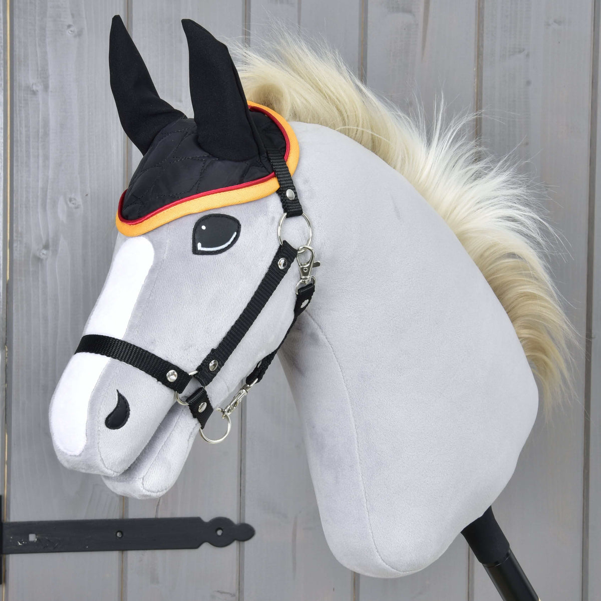 Szőke sörényű, szürke Hobby Horse ló fekete kötőfékben, a fekete-piros-arany Flag DE fülvédővel