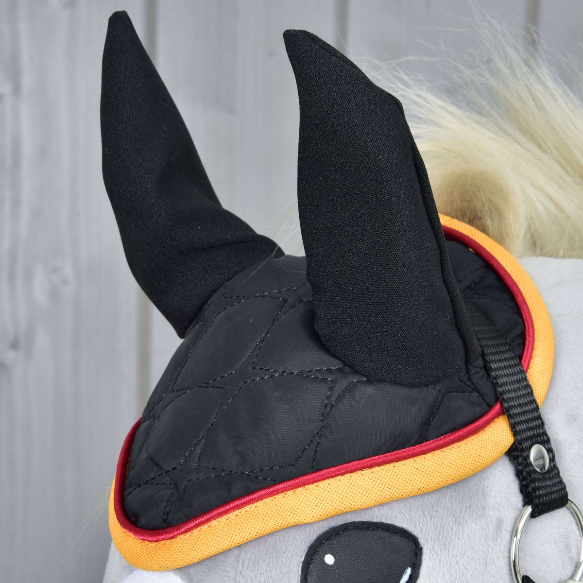 A Flag DE fülvédő közelről egy szürke Hobby Horse lovon: fekete fülek és steppelés piros és aranysárga szegéllyel keretezve