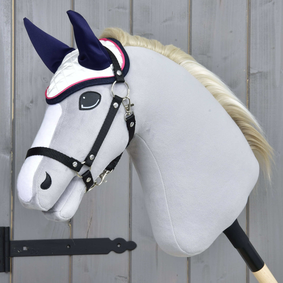 Szürke plüss Hobby Horse ló krémszínű sörénnyel és fekete kötőfékkel, a sötétkék fülrészes, rózsaszín díszítésű fehér fülvédőben