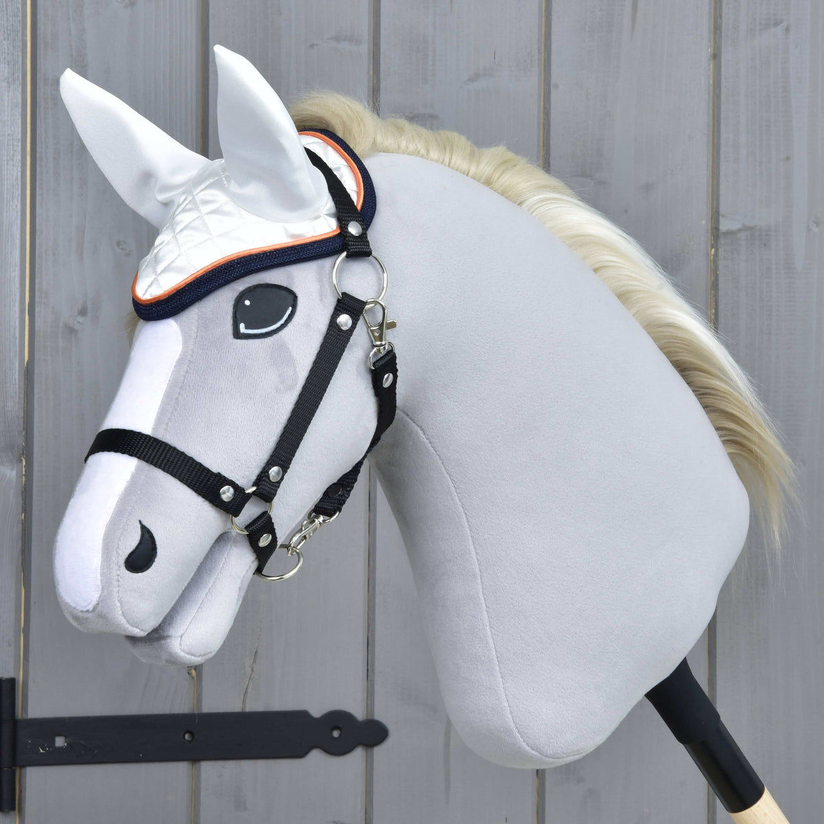 Szürke plüss Hobby Horse ló krémszínű sörénnyel és fekete kötőfékkel, a narancssárga és sötétkék díszítésű fehér fülvédőben