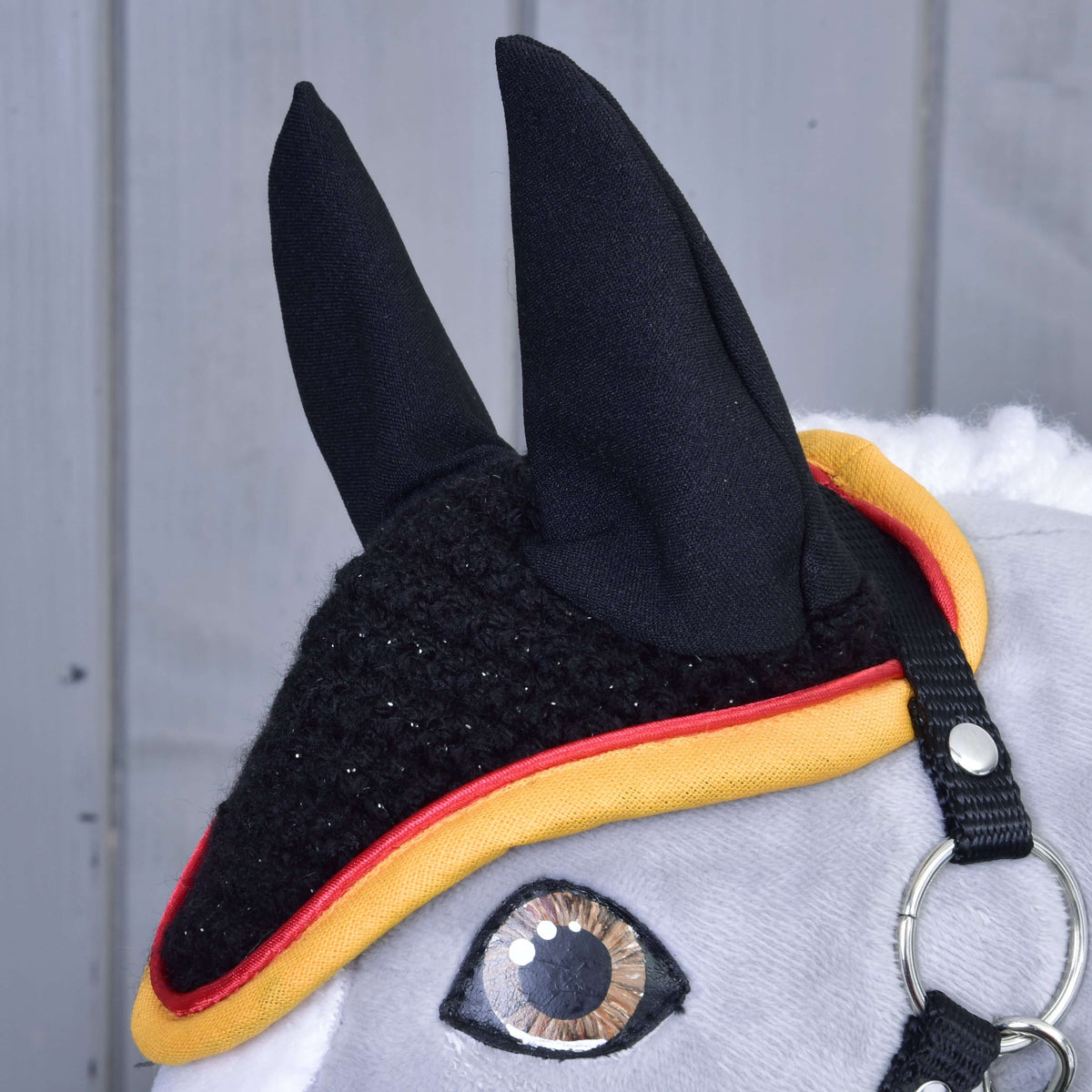 A Flag fülvédő csillogó fekete horgolása, fekete fülei és piros-sárga paszpólja közelről egy Hobby Horse lovon