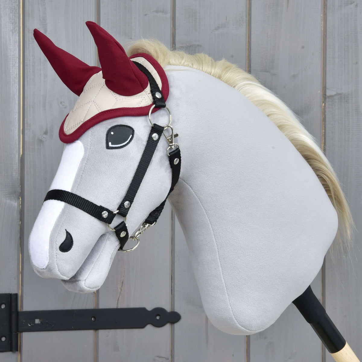 Light grey Hobby Horse with a blond mane wearing the Ear Bonnet Maroon Star and a black halter by a grey wooden door