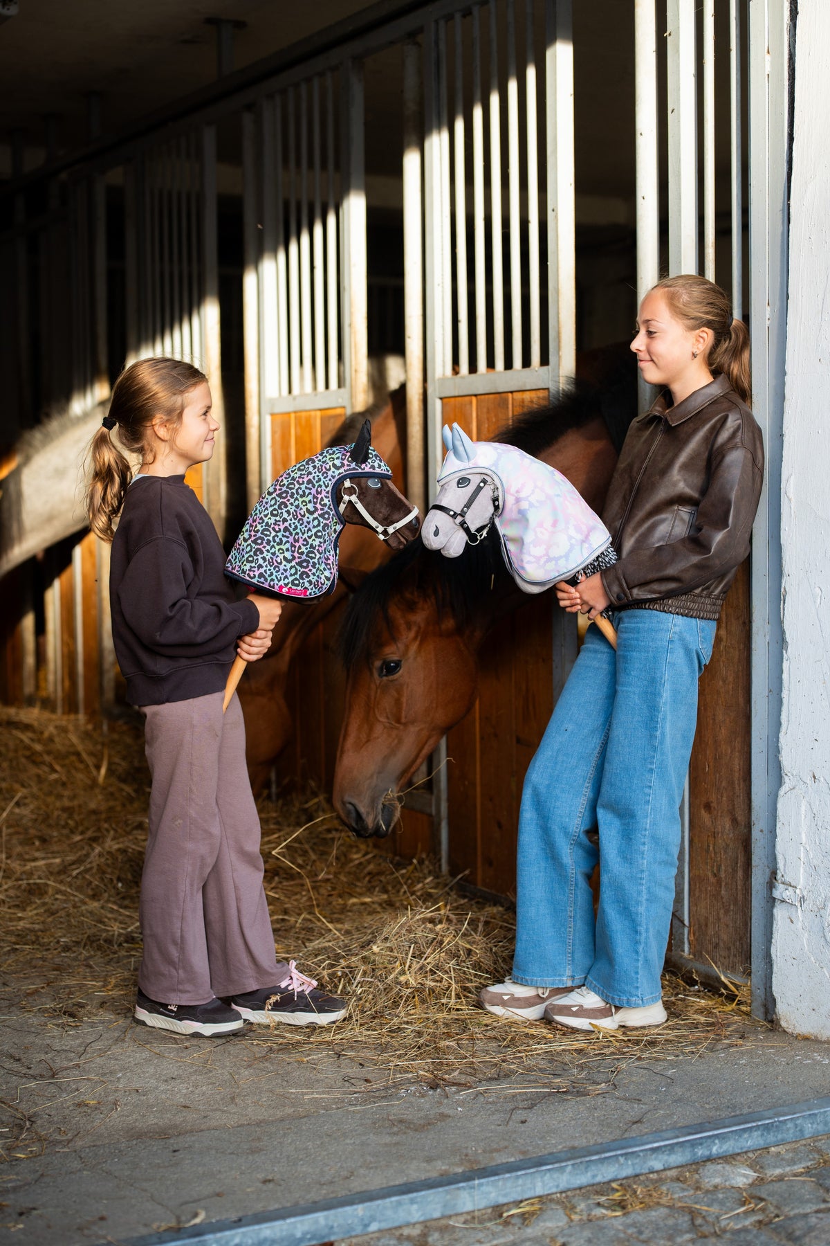 Két lány egy istállóban a Hobby Horse Alfredo lóval és egy szürke Hobby Horse lóval, színes kapucnis esőtakarókban, egy pej ló mellett