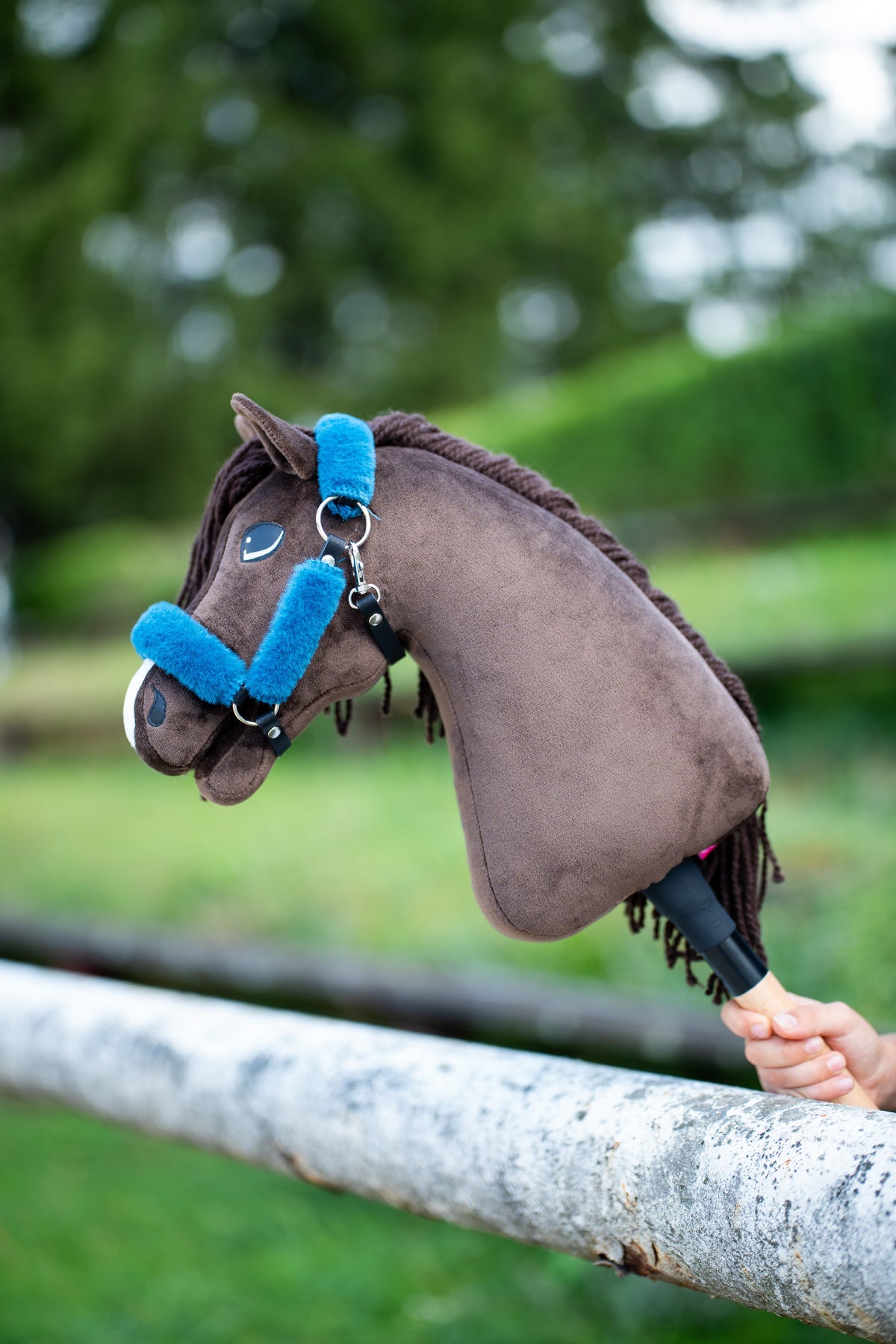 Hobby Horse Chianti petrolkék szőrmés fekete kötőfékben egy fehér nyírfa korláton pihen zöld bokrok előtt