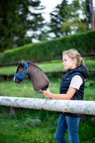 Girl in a navy padded gilet at a white rail holding the dark brown Hobby Horse in a blue fur halter, hedge behind
