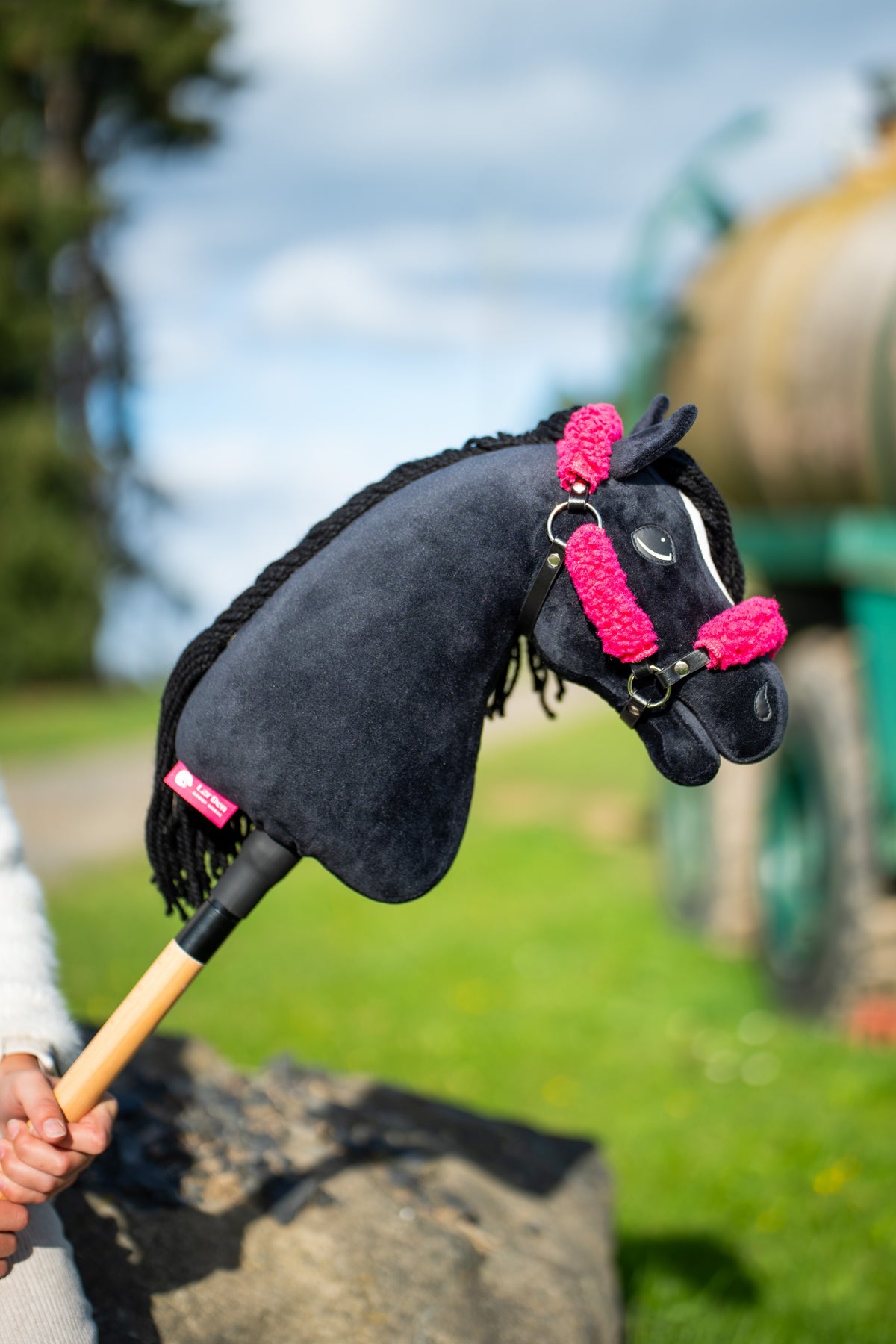 Black plush Hobby Horse with a white stripe and black yarn mane in the black halter with pink fur, held up outdoors