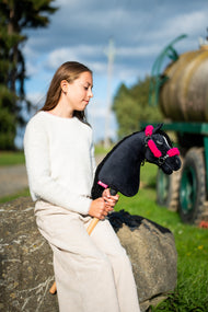 Krémszínű pulóveres lány egy nagy sziklán ül a szabadban egy fekete Hobby Horse lóval, amelyen a rózsaszín szőrmés fekete kötőfék van