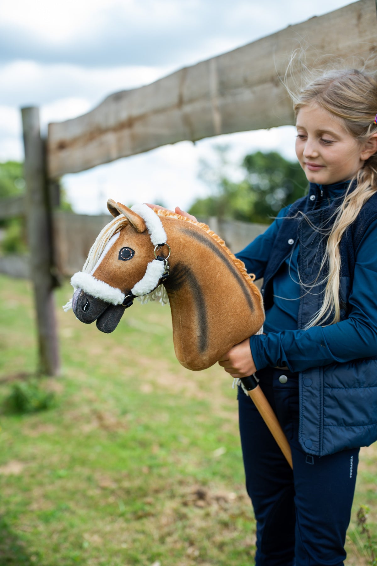 Sötétkék mellényes lány egy karám kerítésénél sötét csíkos, világosbarna Hobby Horse lovat tart a fehér szőrmés kötőfékben