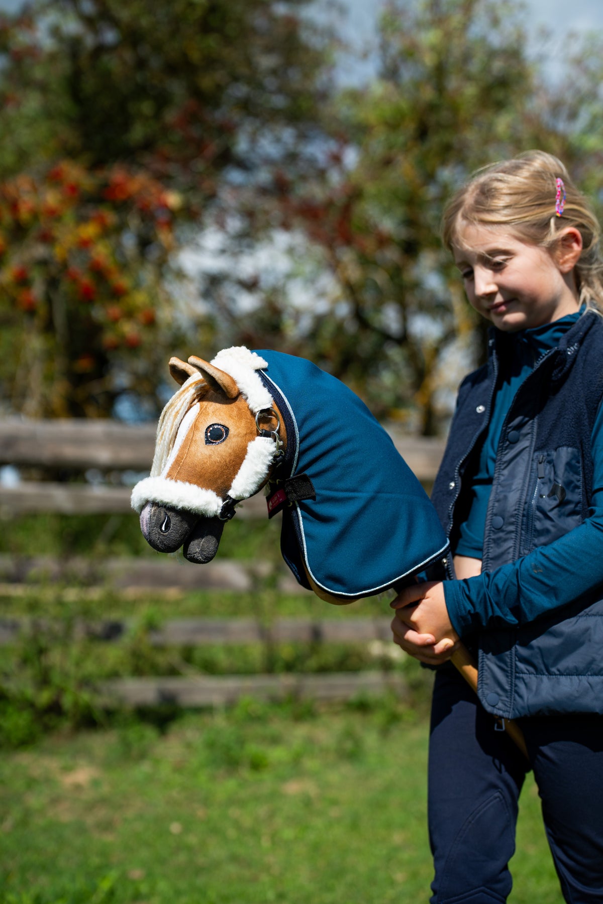 Lány az aranyszínű Hobby Horse lóval, amelyen petrolkék takaró és fehér szőrmés kötőfék van, mögötte őszi bogyók