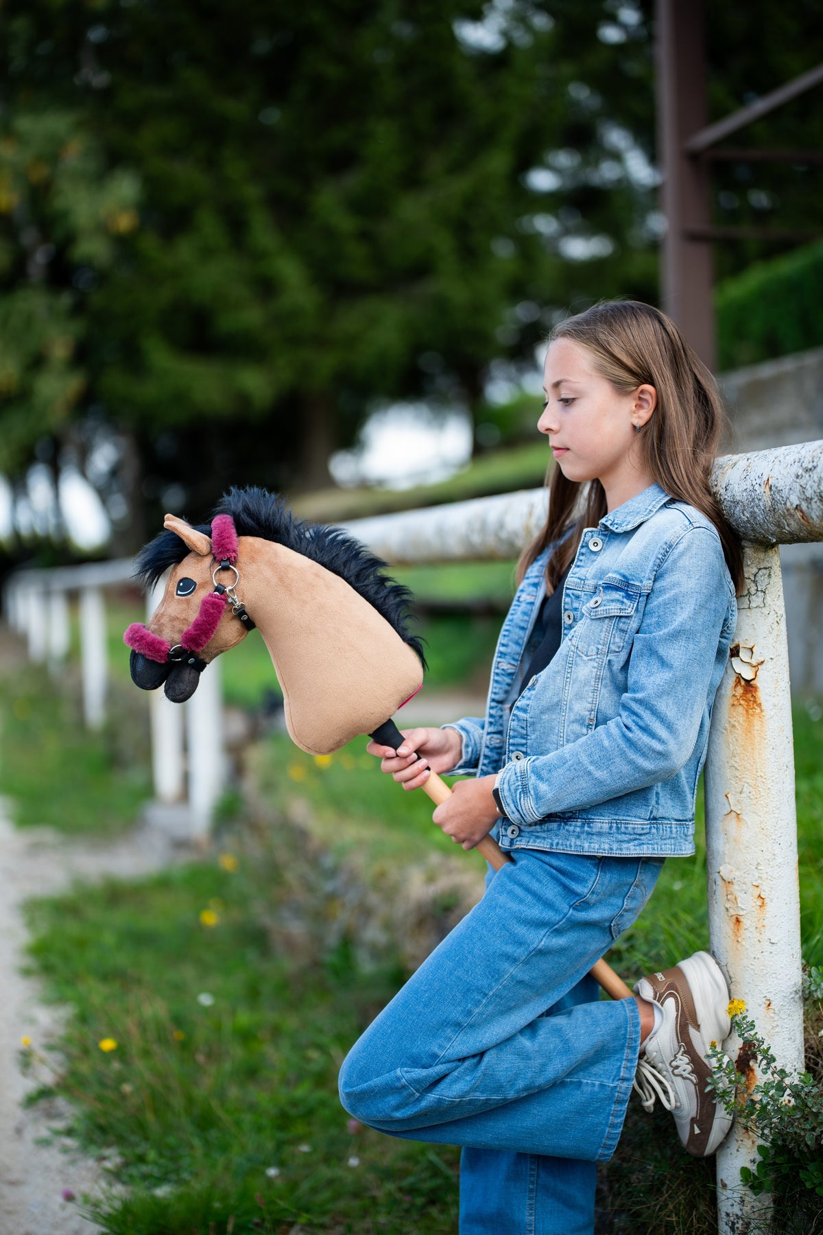 Farmerdzsekis lány egy fehér korlátnak dőlve, kezében a világosbarna Hobby Horse ló málnaszínű szőrmés kötőfékben