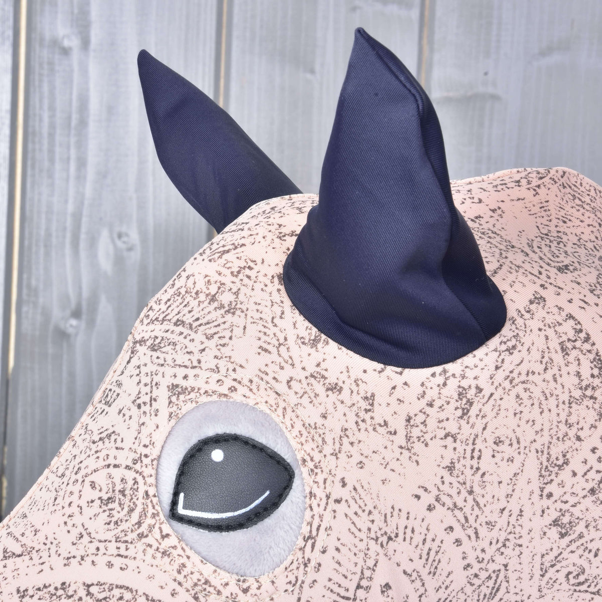 A rózsaszín kasmírmintás Lycra védőruha sötétkék fülei és kerek szemnyílása közelről egy szürke Hobby Horse lovon