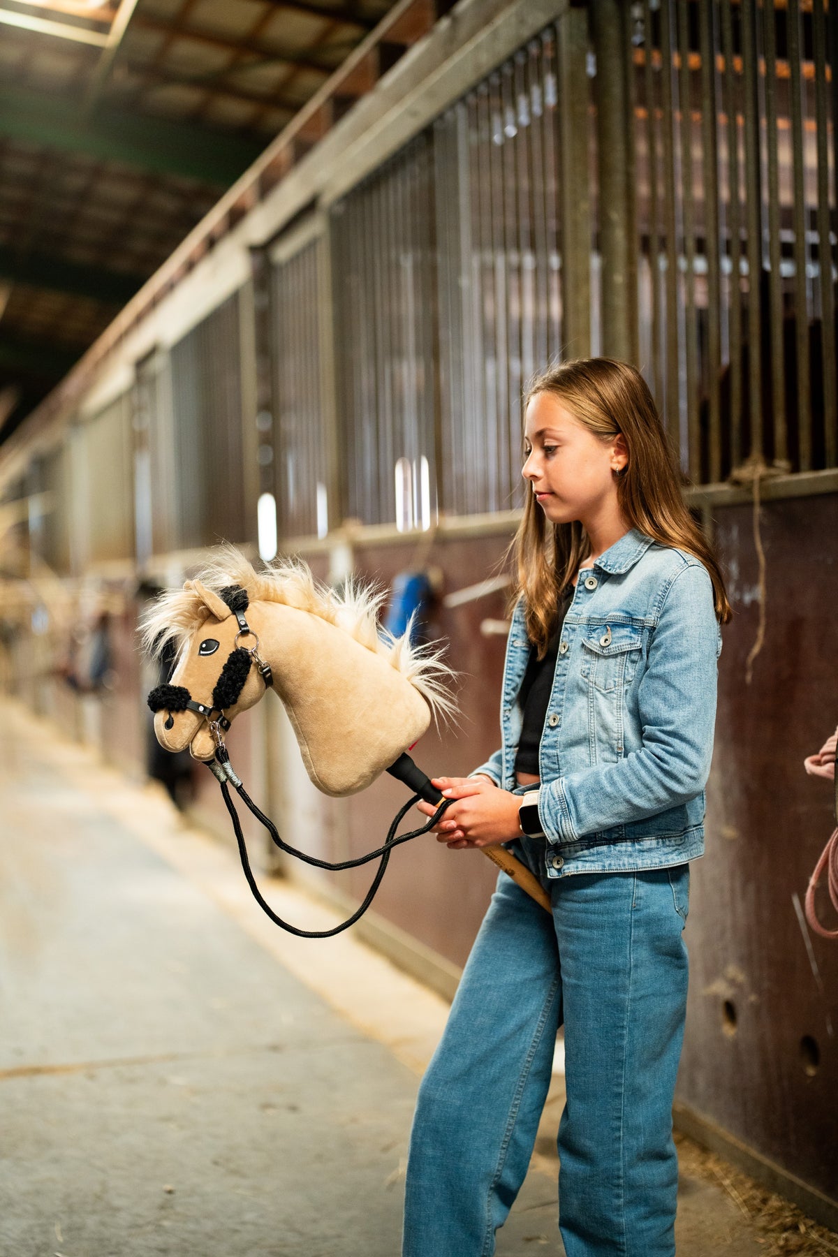 Girl in a denim jacket and jeans holds a cream Hobby Horse with black fur halter on the black reins in a stable aisle