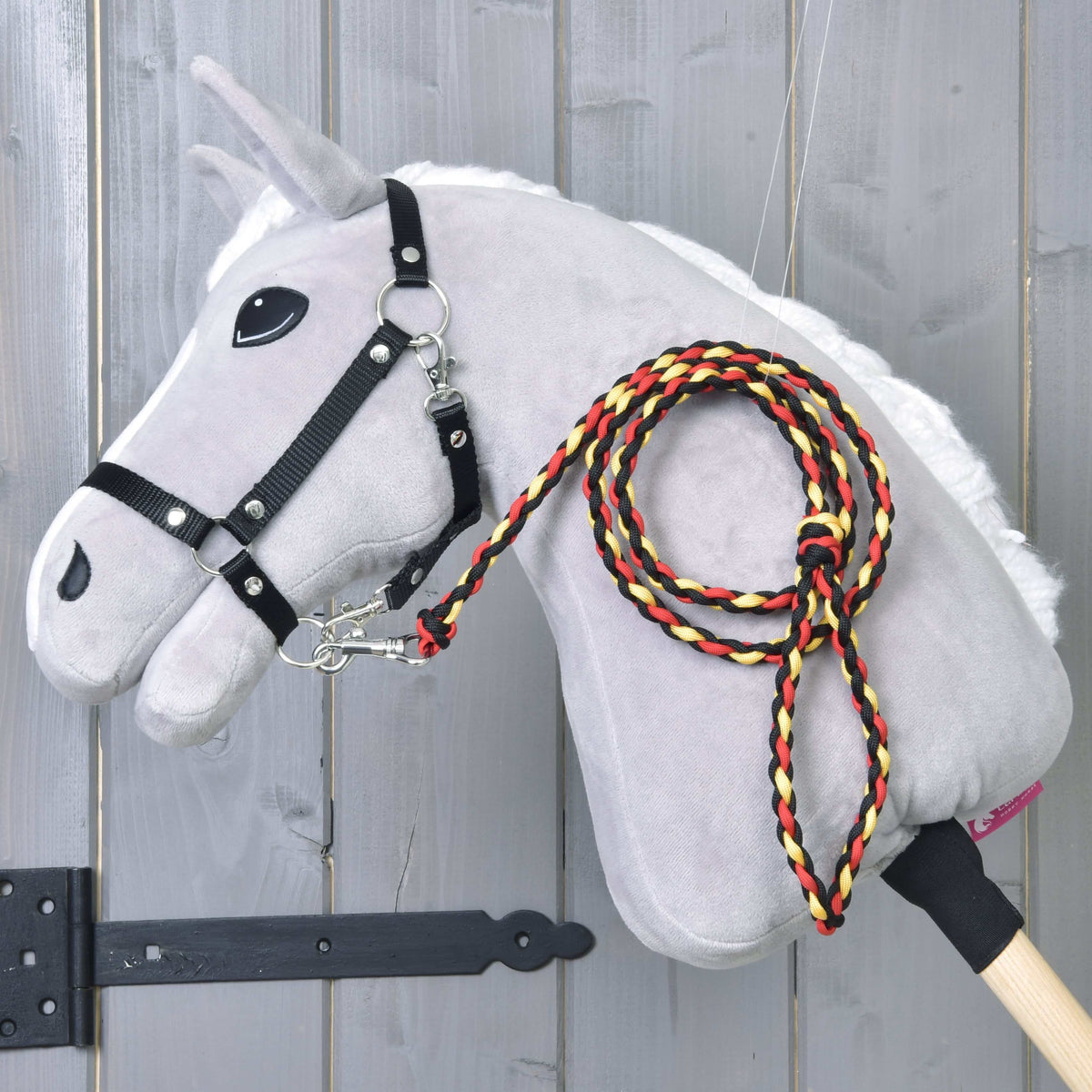 Világosszürke Hobby Horse ló fekete kötőfékben, a fekete-piros-sárga fonott Flag csíkos vezetőszárral