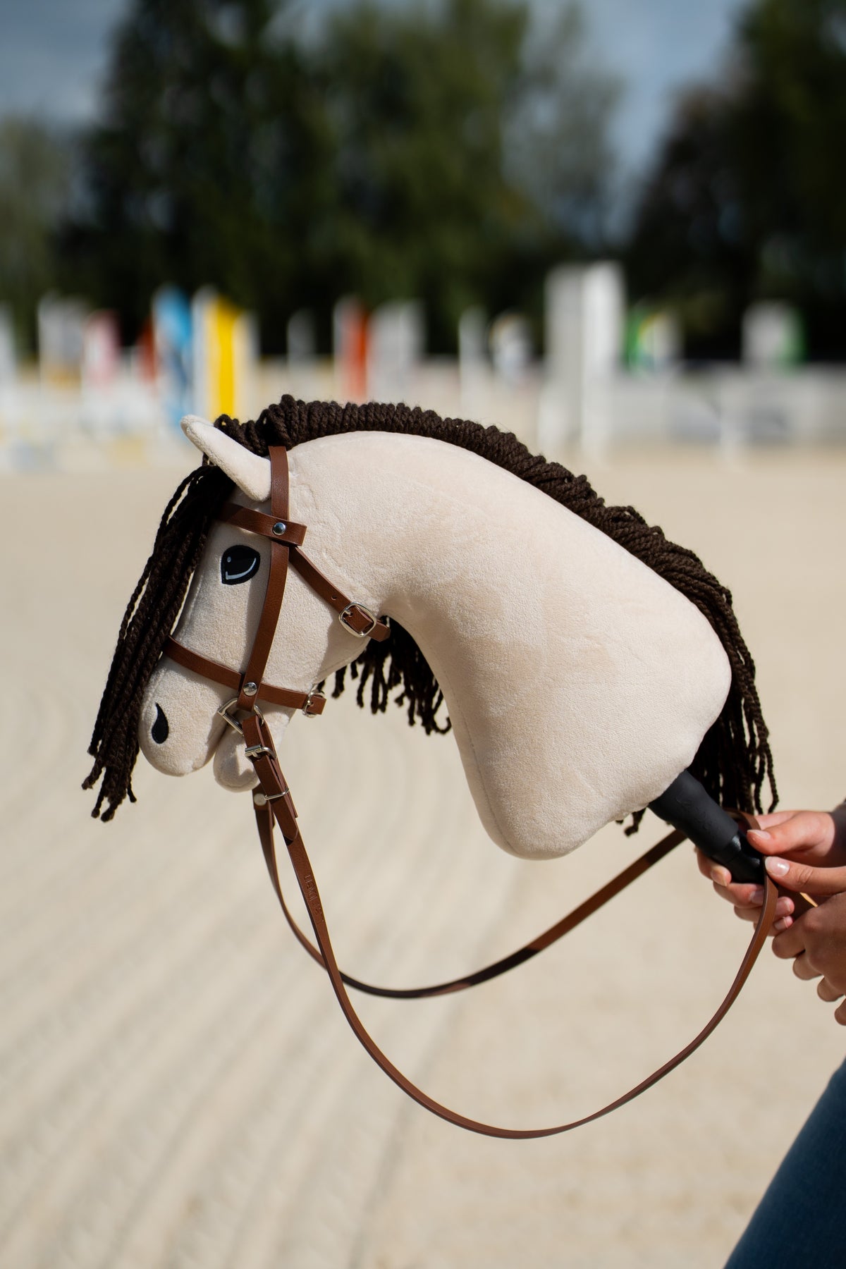 Krémszínű Hobby Horse Frederik LD sötétbarna fonalsörénnyel, orrszíjas barna kantárban, hosszú szárral, mögötte akadályok