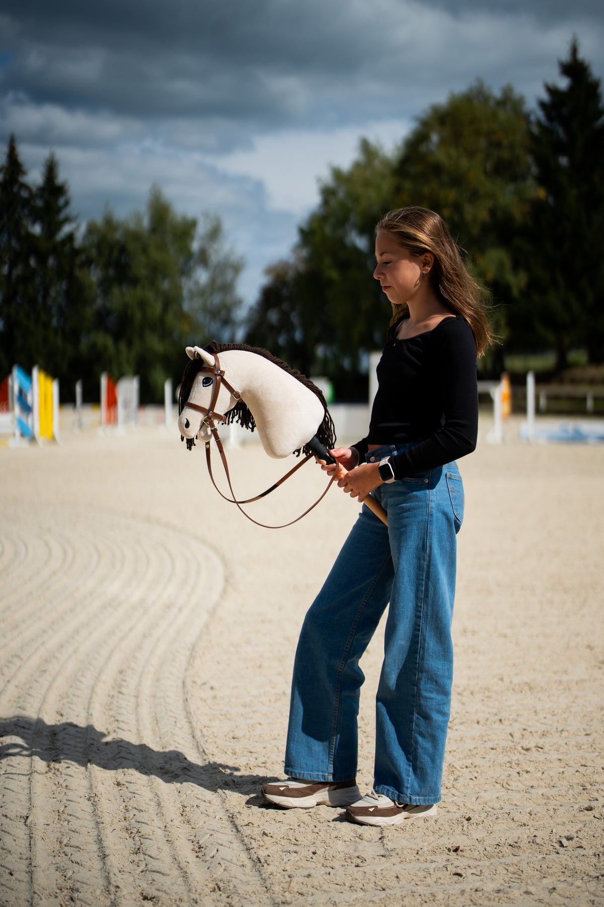 Lány homokos pályán színes akadályok között, kezében krémszínű Hobby Horse ló a barna, orrszíjas komplett kantárban