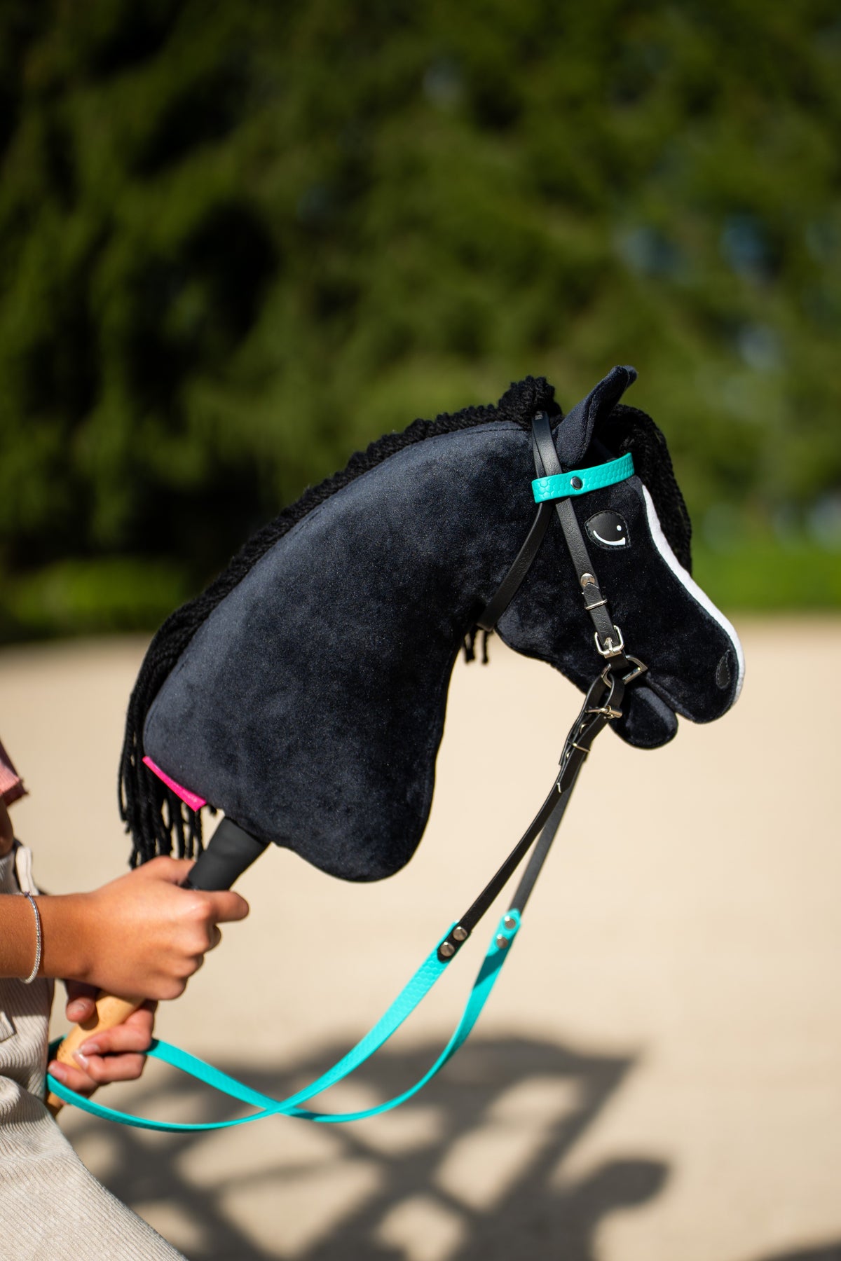 Hobby Horse Freya türkiz homlokszíjas fekete kantárban, türkiz szárral, a magasba tartva egy napsütéses homokos pályán