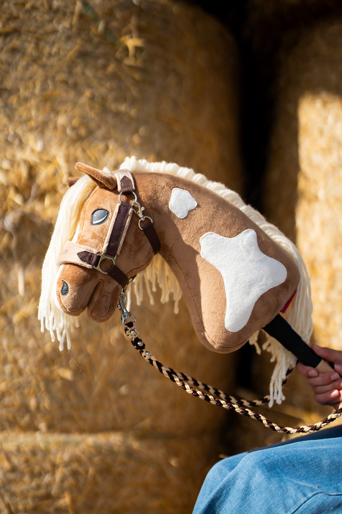 Fehér foltos Hobby Horse Fury barna kötőfékben, barna-krémszínű fonott vezetőszárral, mögötte szalmabálák