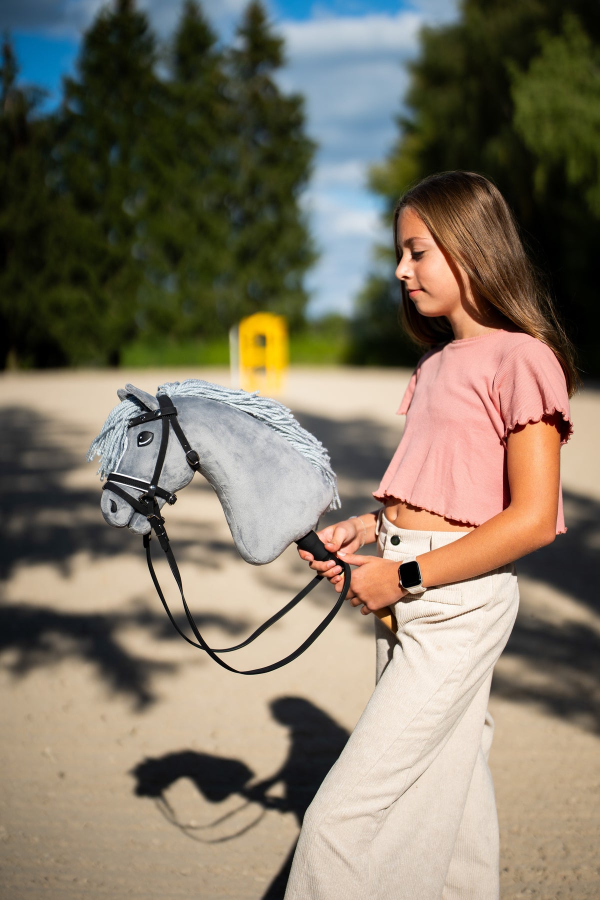 Rózsaszín felsős lány sétál át egy homokos pályán a Hobby Horse Grace lóval, fekete kantárban, szárral