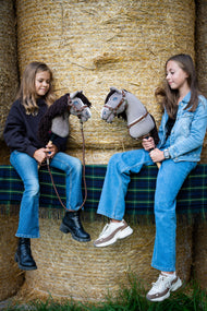 Two girls on straw bales covered with a green tartan throw, each holding a grey Hobby Horse in a brown halter