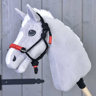 Szürke plüss Hobby Horse ló fehér sörénnyel, a fekete-piros csomózott kötőfékben, szürke fafal előtt