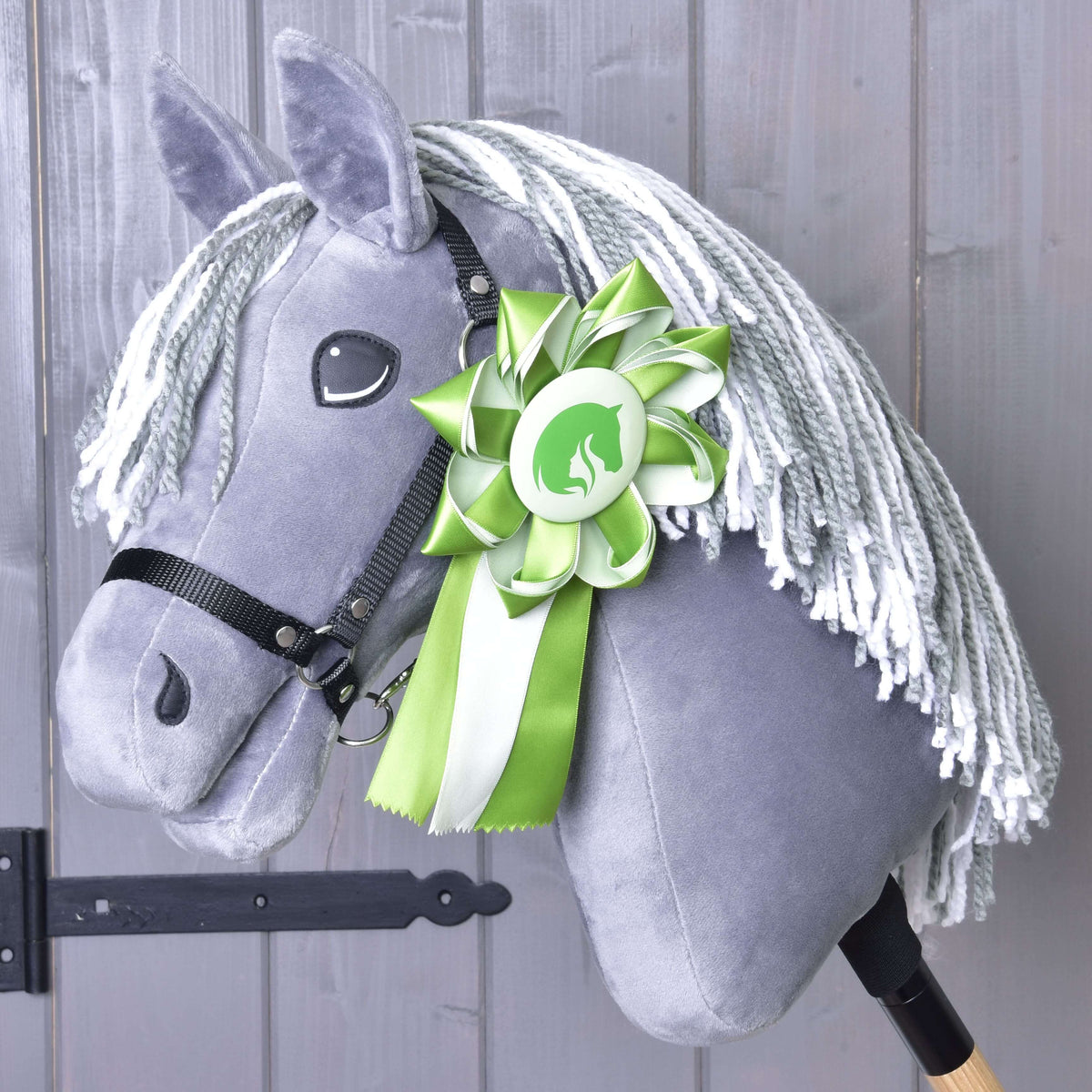 Szürke plüss Hobby Horse ló szürke-fehér fonalsörénnyel és fekete kötőfékkel, a nyakára tűzve a zöld rozetta