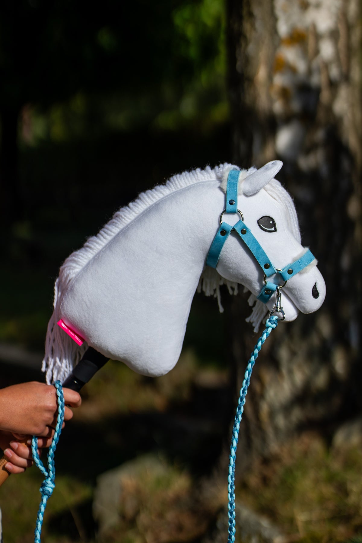 Fehér Hobby Horse Lady közelről pasztellkék kötőfékben, rácsatolt világoskék fonott vezetőszárral, mögötte nyírfatörzs