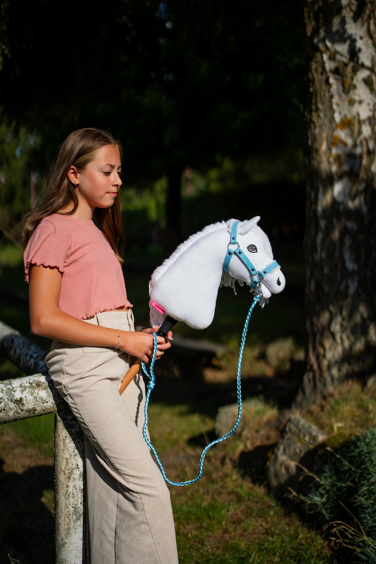 Rózsaszín felsős lány egy nyírfa mellett, kezében pasztellkék kötőfékes fehér Hobby Horse ló, a vezetőszár hurokban lóg