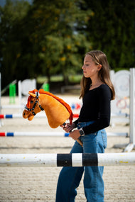 Fekete felsős lány homokos pályán díjugrató akadályok között, kezében egy gesztenyebarna Hobby Horse ló fekete, fehér szőrmés kötőfékben