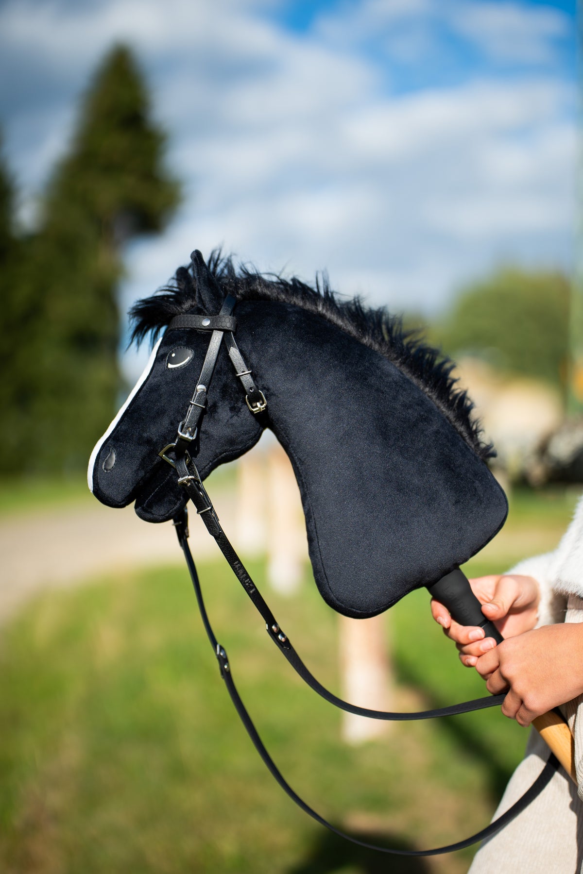 Hobby Horse Loki bozontos fekete sörénnyel, fekete műbőr kantárban, hosszú szárral, mögötte kék ég és egy fenyő