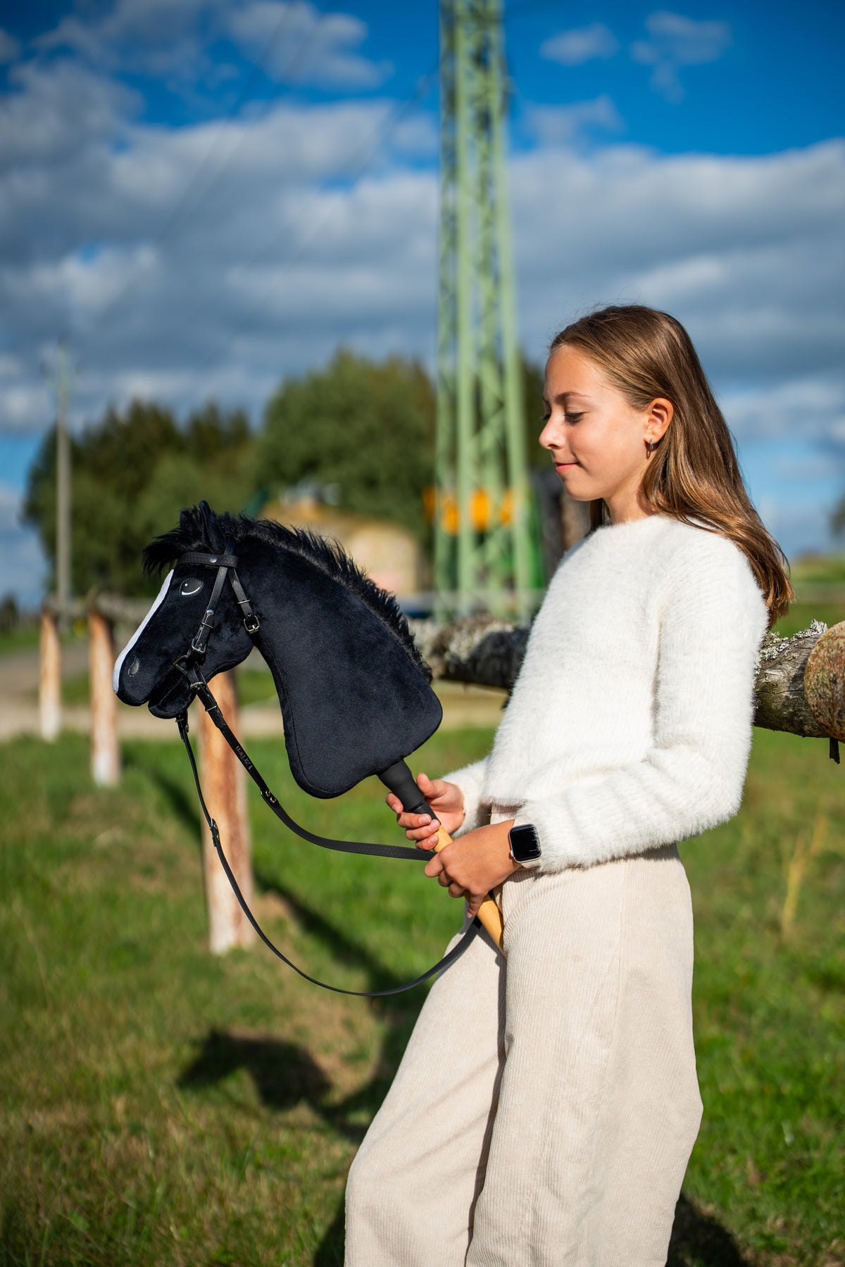 Bolyhos fehér pulóveres lány egy napsütötte réten, kezében a Hobby Horse Loki ló fekete kantárban, szárral