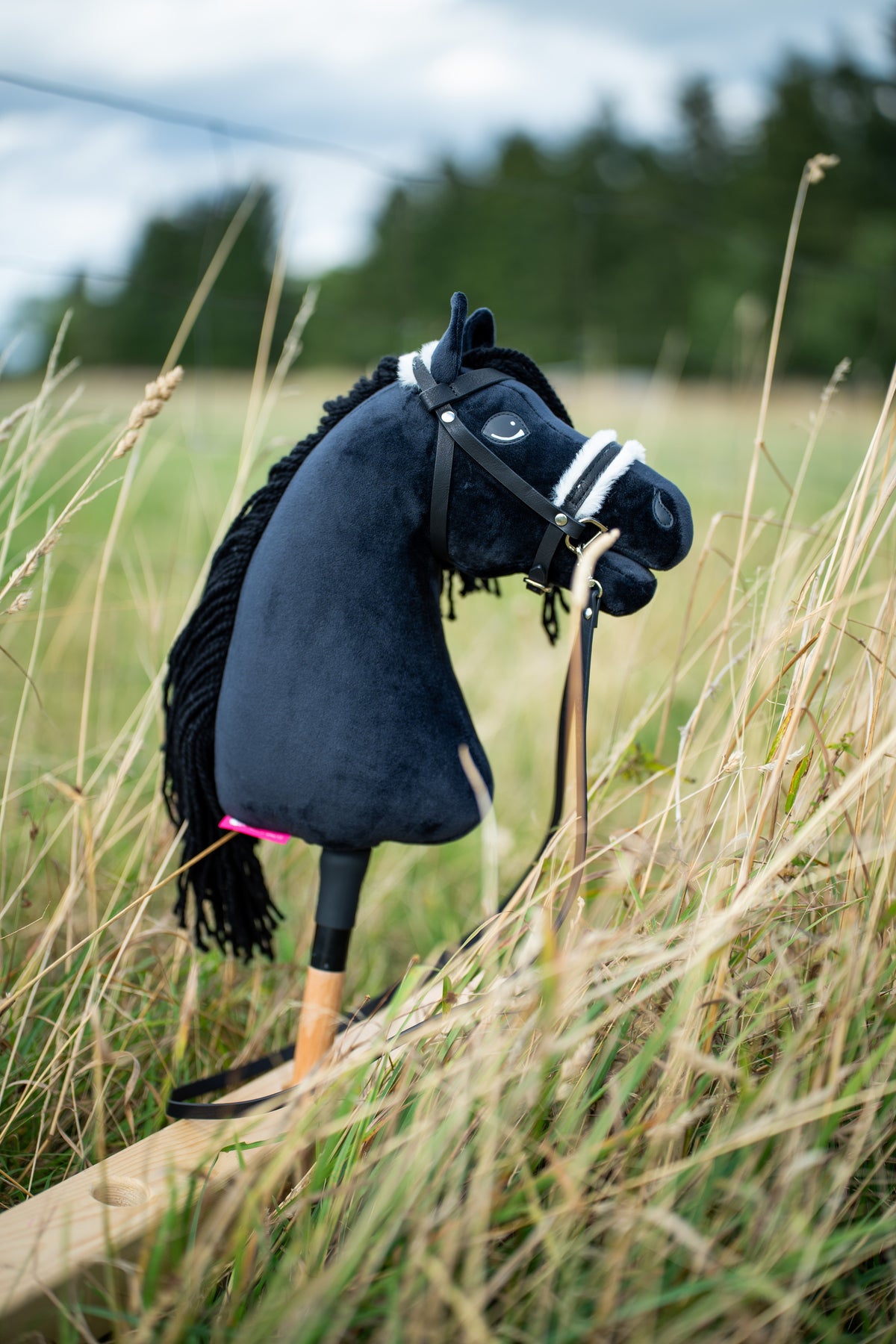 A fekete kötőfékkel kapható Hobby Horse Mahal ló magas réti fűben áll, itt fekete kantárral és fehér szőrmés orrszíjjal