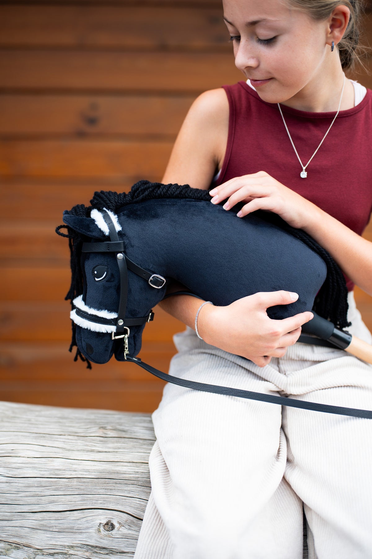 Bordó felsős lány a karjában tartja a fekete kötőfékkel kapható Hobby Horse Mahal lovat, itt fekete kantárral és fehér szőrmével